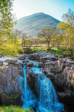 View of Steall Waterfall cascading over rugged rocks into a pool, surrounded by lush green trees and a mountain backdrop under bright sunlight in Fort William, Scotland, United Kingdom.