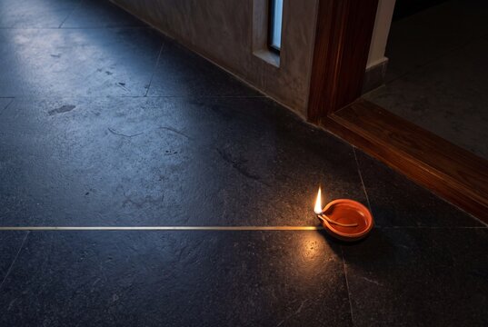 A single traditional clay oil lamp burning brightly on a dark slate floor next to a wooden doorway threshold during an Indian festival celebration