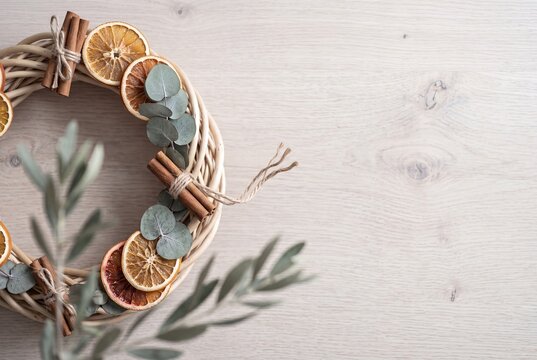 Handmade rustic wicker wreath decorated with dried orange slices, cinnamon sticks tied with twine, and fresh eucalyptus leaves on a light wooden background with copy space