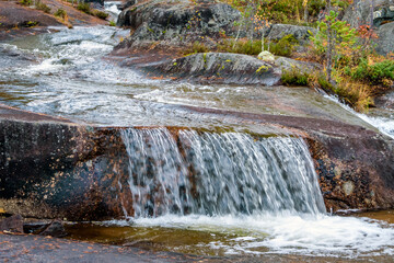 Clear water cascades over smooth layered rocks into a small waterfall at Storforsen, framed by vibrant autumn foliage and mossy stones in a serene Swedish river landscape © Alexandre Patchine