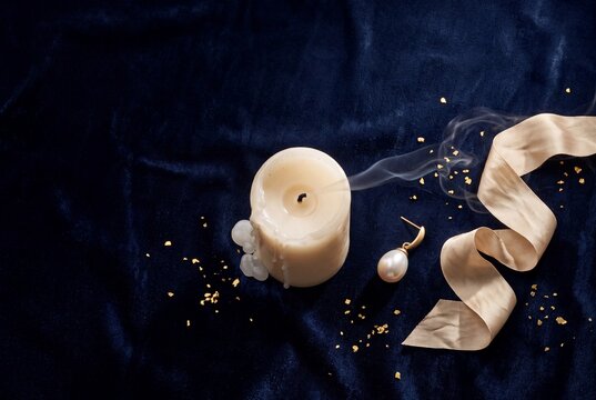 Extinguished white pillar candle with rising smoke, golden ribbon, pearl earring, and gold leaf flakes on dark blue velvet background