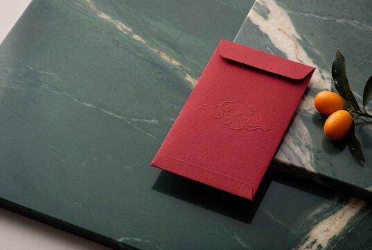 Red envelope with embossed cloud motif resting on dark green marble slabs beside fresh kumquats with leaves, creating a festive and elegant Lunar New Year flat lay composition