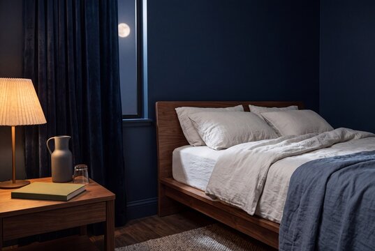 Cozy modern bedroom interior at night with dark blue walls, wooden bed frame, linen bedding, and a glowing bedside lamp