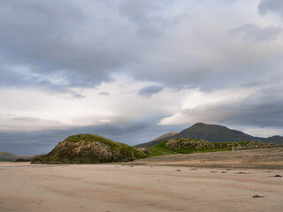 Bare sandy beach stretches toward grassy, rounded hills under cloudy sky. Hills rise gently with patches of green and exposed rock. Scene feels quiet and serene, perfect for nature or travel themes © mark_gusev