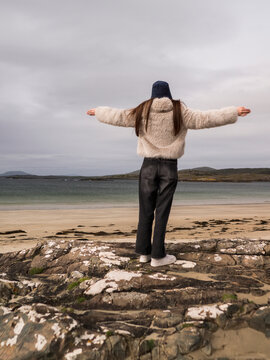 Young woman stands on sandy beach with waves crashing behind her, hands up. She wears fluffy coat and dark jeans, looking directly at camera. Her expression is calm and confident under overcast sky