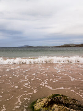 Pink-hued seafoam crests near a mossy rock on a calm beach. Waves gently roll toward shore under an overcast sky. Distant hills fade into soft mist on horizon