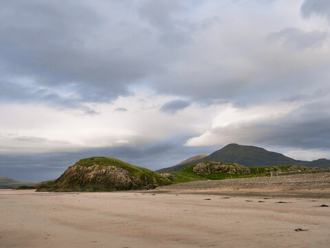 Bare sandy beach stretches toward grassy, rounded hills under cloudy sky. Hills rise gently with patches of green and exposed rock. Scene feels quiet and serene, perfect for nature or travel themes