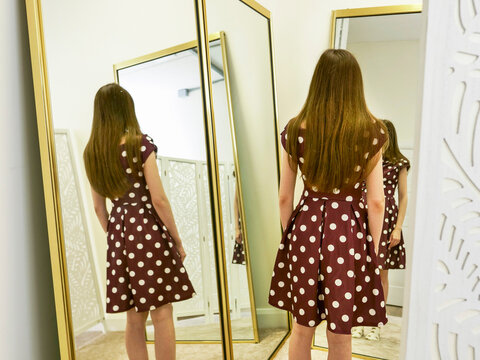 Young girl with long hair stands before tall, golden-framed mirrors. She wears a polka-dot dress, examining her fashion stylish outfit. Her posture suggests quiet curiosity and self-discovery