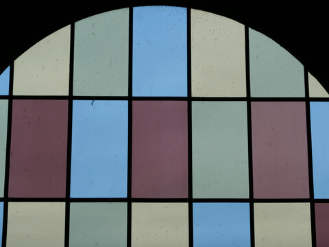 top arched portion of a stained glass window, divided into a grid of pastel blue, grey, purple, and cream panes by black frames