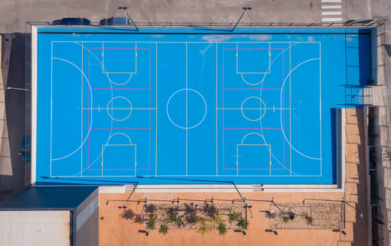 Overhead view of a bright blue outdoor multi-sport court with white markings and purple key lines beside a brick building with planted trees