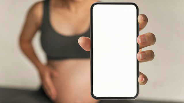 Expecting Mother Holding Smartphone With Blank Screen In Front Of Pregnant Belly, Standing In Cozy Home Environment. Anticipation And Joy Of New Life. Maternity Concept.