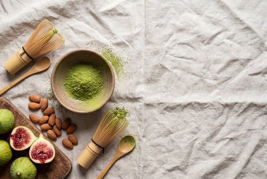 Top view of vibrant green matcha tea powder in a ceramic bowl with bamboo whisks, wooden spoons, almonds, and fresh figs on a crumpled linen cloth with copy space