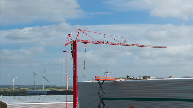 Aerial view of a red tower crane at an industrial warehouse construction site with workers on the roof and wind turbines in the distance in Daventry, England, United Kingdom.