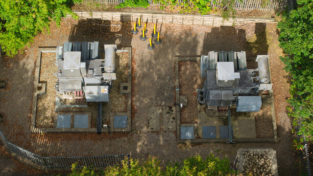 Aerial view of two electrical transformers situated on concrete pads within a fenced enclosure, surrounded by trees and foliage in Lutterworth, England, United Kingdom.