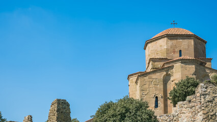 Jvari Monastery stone church with domed tiled roof and cross above weathered ruins and greenery under a clear blue sky in Mtskheta, Georgia © Mila
