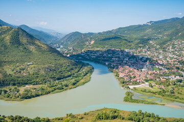 Aerial panorama of Mtskheta, Georgia, showing the Kura and Aragvi river confluence, hillside neighborhoods, green mountains, and a church © Mila