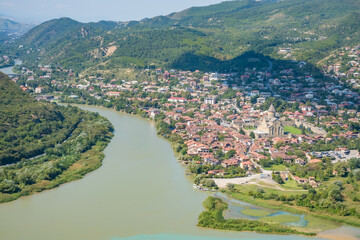 Aerial panorama of Mtskheta, Georgia, with the Kura and Aragvi rivers, historic church, red-roofed homes, wooded hills, and wetlands © Mila