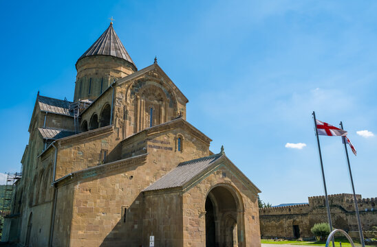 Svetitskhoveli Cathedral in Mtskheta, Georgia, featuring carved stone walls, conical dome, Georgian flags, fortress wall, and blue sky