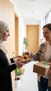 Vertical photo of a woman in a hijab offering baklava to her neighbor in an apartment hallway. Diverse women sharing food and a wrapped gift. Community and hospitality concept