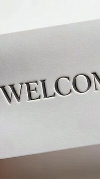 Vertical video: Tilting hand rotating white WELCOME card on table revealing thumb and embossed text