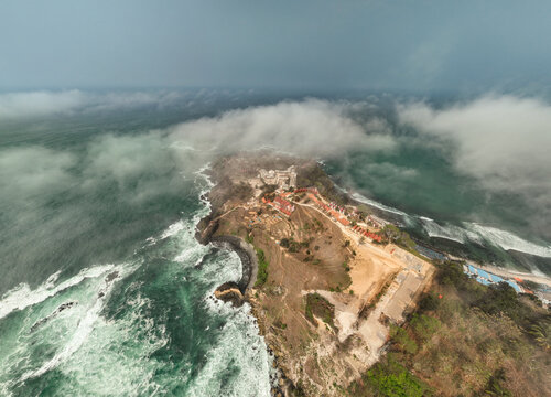 Aerial view of Menganti Beach with rugged cliffs, crashing waves, and coastal buildings under a cloudy sky in Kecamatan Ayah, Jawa Tengah, Indonesia.