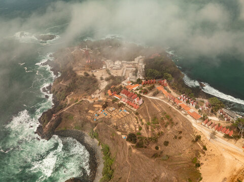 Aerial view of Menganti Beach featuring coastal cliffs, resort buildings with red roofs, and lush vegetation partially obscured by clouds in Kecamatan Ayah, Jawa Tengah, Indonesia.