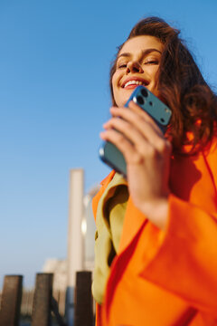 Mobile banking app, smiling young woman in an orange coat holds a smartphone near her face against a blue sky. Concept of online finance, digital payments and convenient money management on the go.