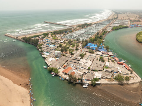Aerial view of Cilacap Shrimp Farm showing aquaculture ponds, coastal buildings, and small fishing boats along the river mouth in Nusawungu, Central Java, Indonesia.