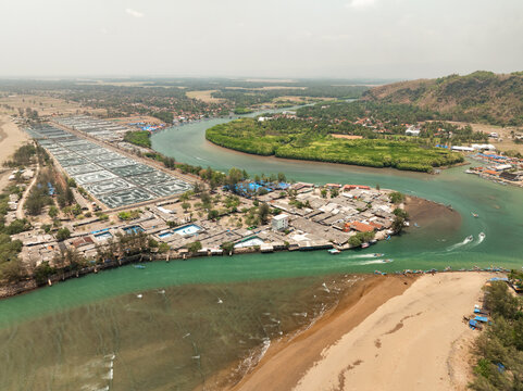 Aerial view of Cilacap Shrimp Farm situated along the river mouth with nearby coastal village, fishing boats, and lush mangrove forests in Nusawungu, Central Java, Indonesia.