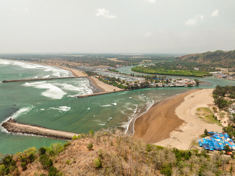 Aerial view of the Cilacap Shrimp Farm area featuring breakwaters extending into the sea, a river mouth, sandy beaches, and coastal vegetation in Nusawungu, Central Java, Indonesia.