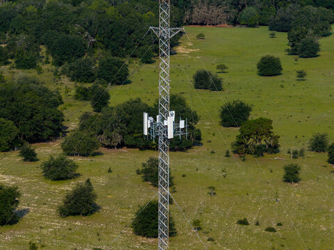 Aerial view of a tall metal cell tower with communication antennas standing in a vast green field surrounded by scattered trees in Old Town, Florida, United States.