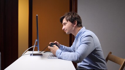 Young man concentrating on a computer screen while playing a video game with a handheld controller at a desk, enjoying digital entertainment during leisure time indoors © Aliaksandr