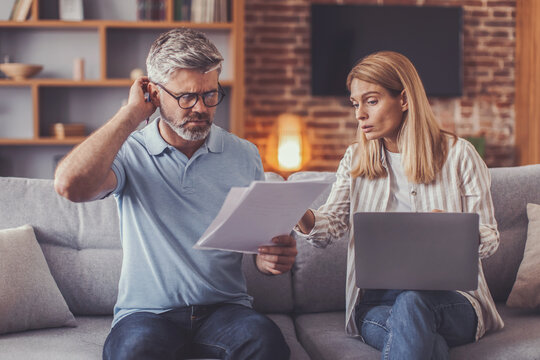Concerned Caucasian Man And Woman Analyzing Papers Together At Home, One Searching For Information On Laptop, Serious Expressions, Engaged In Business Discussion, Living Room Setting, Business Concept