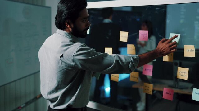 Scrum master explaining task board, bearded man showing sticky notes on glass wall