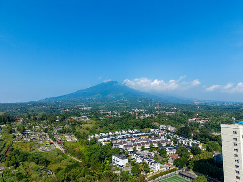 Aerial view of the residential area with Mount Salak in the background, lush green trees, and clear blue sky in Bogor City, West Java, Indonesia.