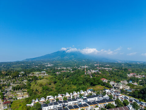 Aerial view of residential houses with modern rooftops nestled in lush greenery with Mount Salak in the background under a clear blue sky in Bogor City, West Java, Indonesia.