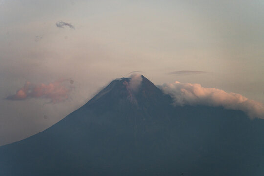 Aerial view of Mount Merapi with volcanic smoke rising from the crater and soft clouds surrounding the peak at sunset in Selo, Central Java, Indonesia.