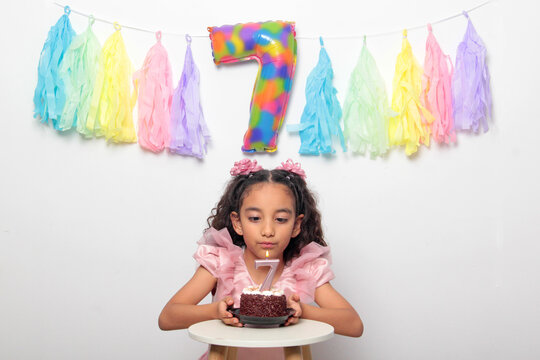 Seven-year-old Latina girl with brown skin and curly hair celebrates her birthday with cake, candle, and colorful decorations