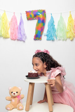 Seven-year-old Latina girl with brown skin and curly hair celebrates her birthday with cake, candle, and colorful decorations
