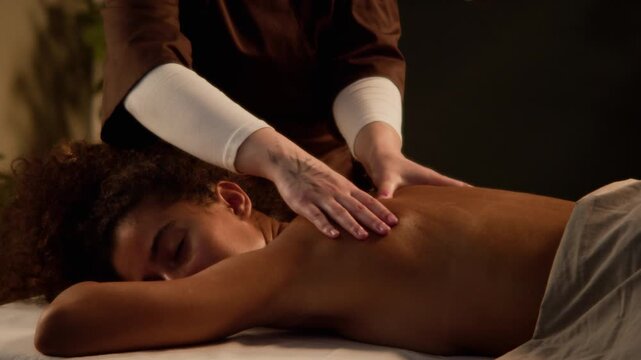 Massage therapist massaging back of relaxed biracial young adult woman lying on treatment table. Footage suitable for spa wellness promotions and body therapy marketing