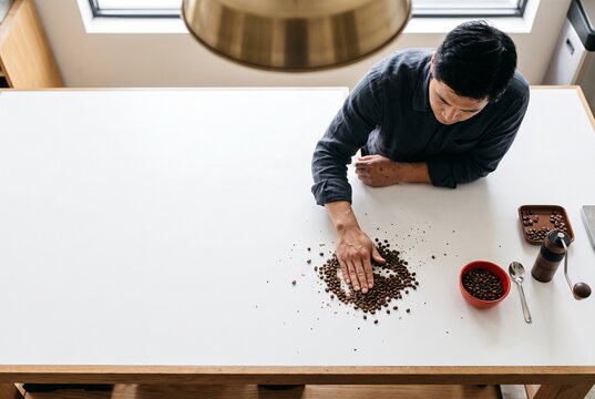 High angle view of a male barista sorting roasted coffee beans by hand on a clean white table with a manual grinder and cupping spoon