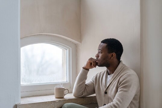Thoughtful young Black man in a beige cardigan sitting by an arched window with a steaming cup of coffee, looking outside in quiet contemplation