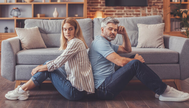 Worried Couple, White Male And Female, Sitting Back To Back On Floor In Living Room, Not Communicating, Reflecting Relationship Strain And Emotional Distance. Home Environment, Unrecognized.