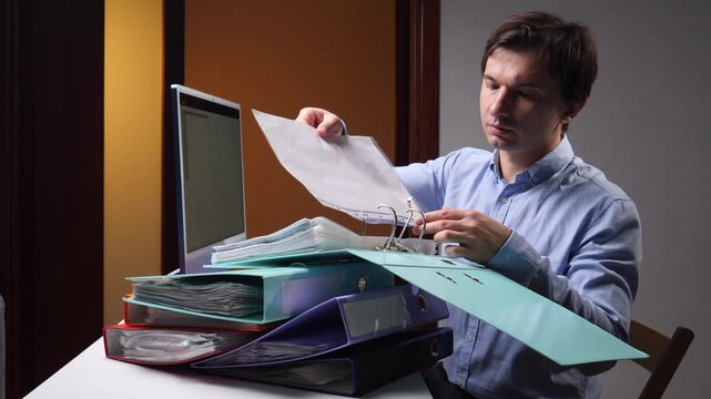 Office worker reviewing paperwork at his desk, examining a sheet in an open ring binder beside stacked folders and a computer, conveying document management and administrative work