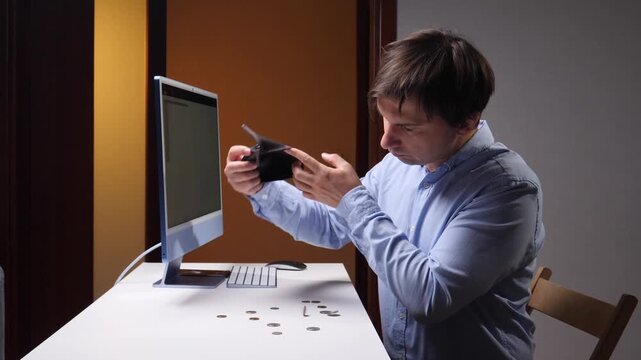 Man inspecting an empty wallet beside a desktop computer, with a few coins scattered across the desk, conveying financial hardship, money worries, debt, and personal budget strain at home