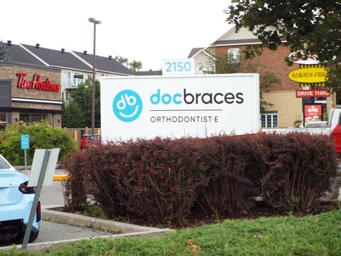 Gloucester, Ontario, Canada &ndash; August 04, 2025: A "Docbraces orthodontist" sign stands behind reddish shrubs with Tim Hortons in the background along a commercial lot streetscape on Montr&eacute;al Rd.
