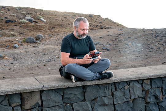 A drone pilot sits cross legged on a low stone wall concentrating on his remote controller near Los Chochos. A candid look at how an aerial shot actually gets made.