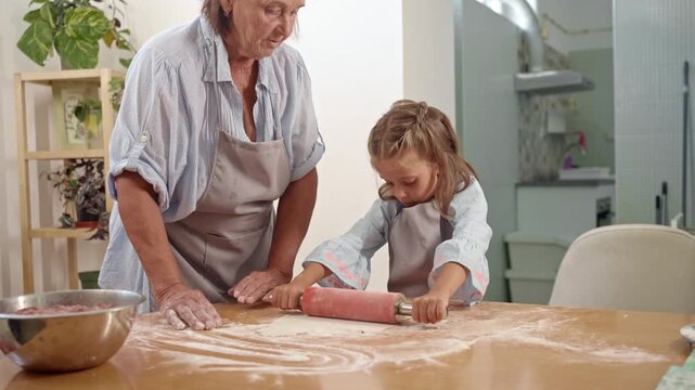 A child rolls out dough on a table while a grandparent watches closely in a bright kitchen. Flour is scattered around as they share a special cooking moment.