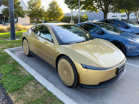 Dallas, Texas, USA - October 15th, 2026: Gold Tesla Cybercab prototype parked next to blue Tesla Model 3 in parking lot outside service center