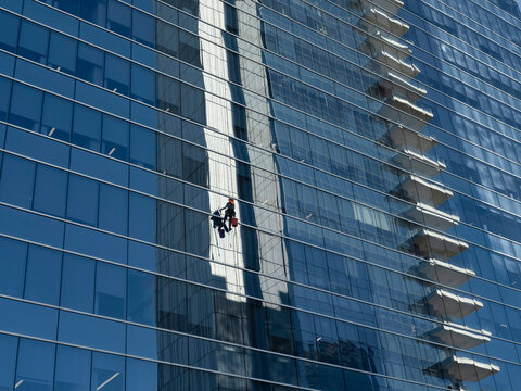 Window washer suspended on ropes while cleaning the glass curtain wall of an office skyscraper in downtown Dallas, with a reflection of another building in the glass.